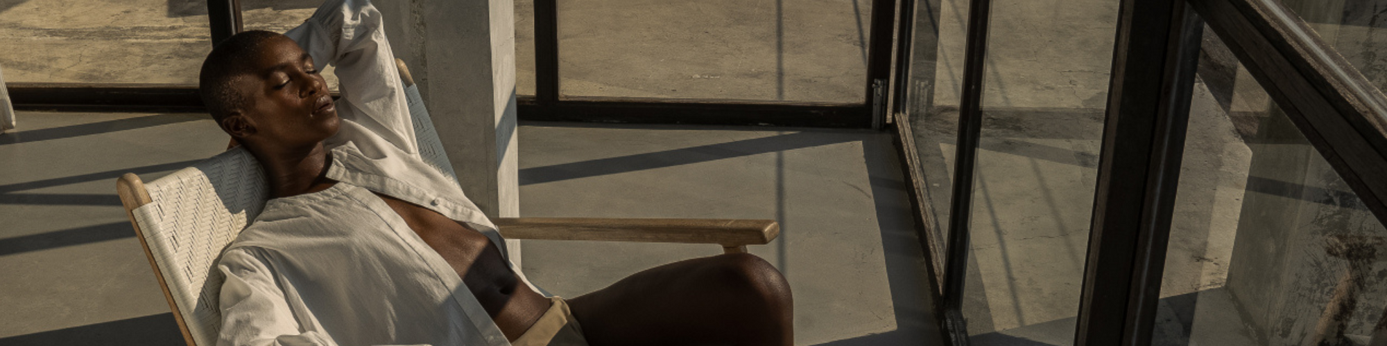 A classically dressed woman in a sustainable white button down and khaki bikini bottoms embodies a life well lived, lounging in a chair in front of sun-drenched windows.