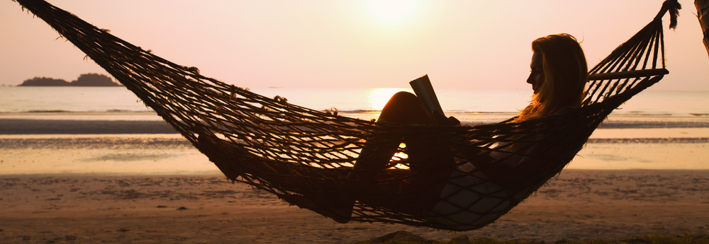 A woman relaxing and reading in a hammock on a beach with the sun setting behind her, demonstrating living well and living in harmony with nature.