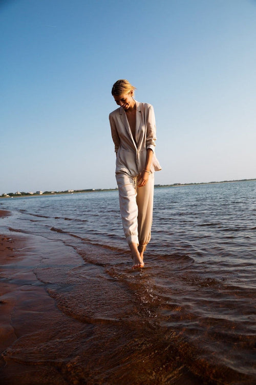 A person wearing eco-friendly Neu Nomads Allie Ankle Pants with an elasticized waistband walks barefoot along the shoreline on a sandy beach during a sunny day.