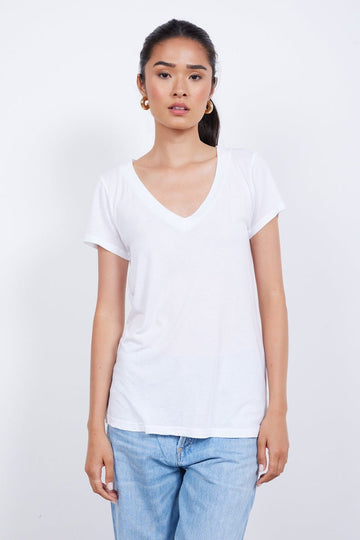 A person with long hair tied back is wearing a plain white, ethically made LOW v NECK BOYFRIEND TEE by LAmade and blue jeans, standing against a plain white background.