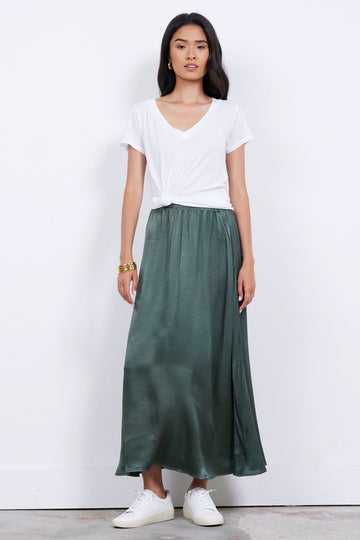 A person stands against a plain background, wearing an ethically made white LOW v NECK BOYFRIEND TEE from LAmade knotted in the front, a long green skirt, white sneakers, and a gold bracelet.