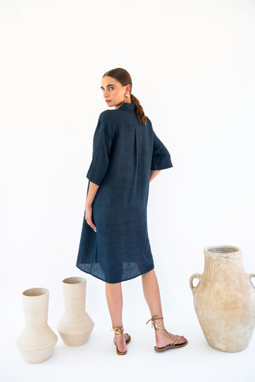 A woman in a dark blue, knee-length shirt dress stands facing backward with her head turned to the camera. She wears strappy sandals and is near three large ceramic vases placed on the floor. The stylish ensemble, a Chloe Shirt Dress - Linen from Neu Nomads, exudes effortless elegance.