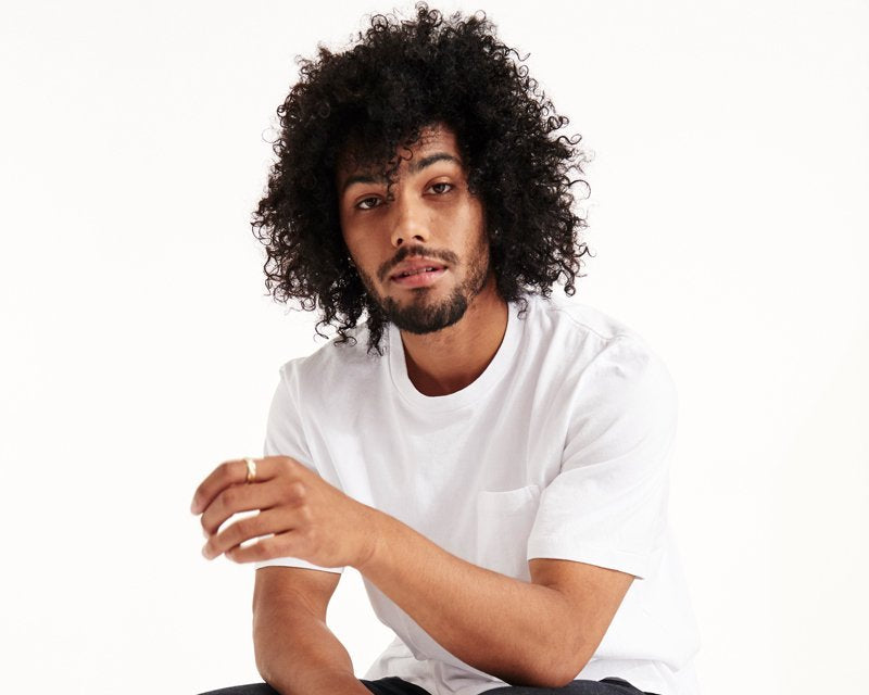 A man with curly hair, wearing an LAmade ECHO PARK TEE W/ POCKET- WHITE with a boxy fit, dark cuffed jeans, and light green sneakers, sits on a wooden stool against a white background.