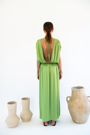 A person stands with their back to the camera, wearing a green backless Natalia Dress - Satin Tencel from Neu Nomads. Three ceramic vases are placed on the floor to their left, making for a refined setting suited for any special occasion.