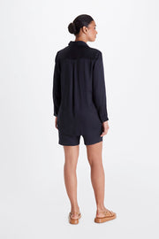 A woman stands facing away, wearing a long-sleeved black Twiggy Boyfriend Shirt by Neu Nomads, black shorts, and tan sandals. Her hair is tied in a bun, and the background is plain white.