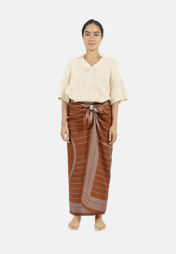 A person stands against a plain background wearing a cream-colored top and an Antibes Beach Sarong TAUPE from 1 People tied at the waist. They have bare feet and a neutral expression, embodying the effortless elegance of this versatile accessory.