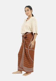 A barefoot woman wearing a loose-knit cream sweater and a rust-colored TENCEL™ Modal wrap skirt stands against a plain white background. She is looking slightly to the side with a neutral expression on her face, showcasing the Antibes Beach Sarong TAUPE from 1 People that can also be styled as a versatile accessory.