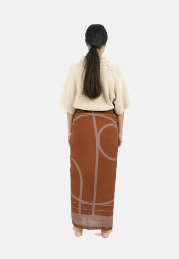 A person with long hair in a ponytail is standing with their back to the camera. They are wearing a beige short-sleeved sweater made of TENCEL™ Modal and an Antibes Beach Sarong TAUPE from 1 People, perfect as a versatile accessory.